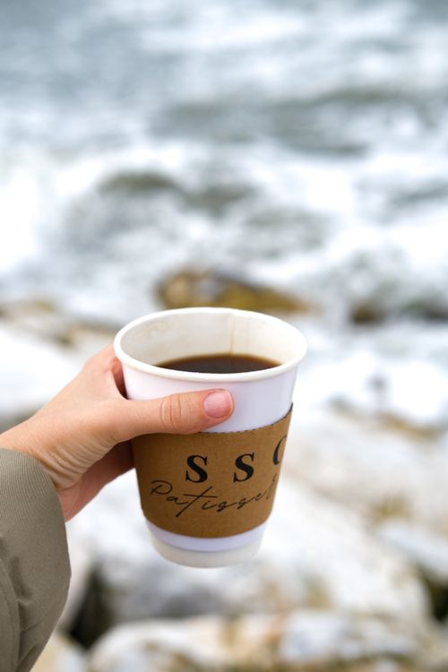 Free Cozy moment capturing a hand holding a coffee cup close to the ocean waves. Stock Photo