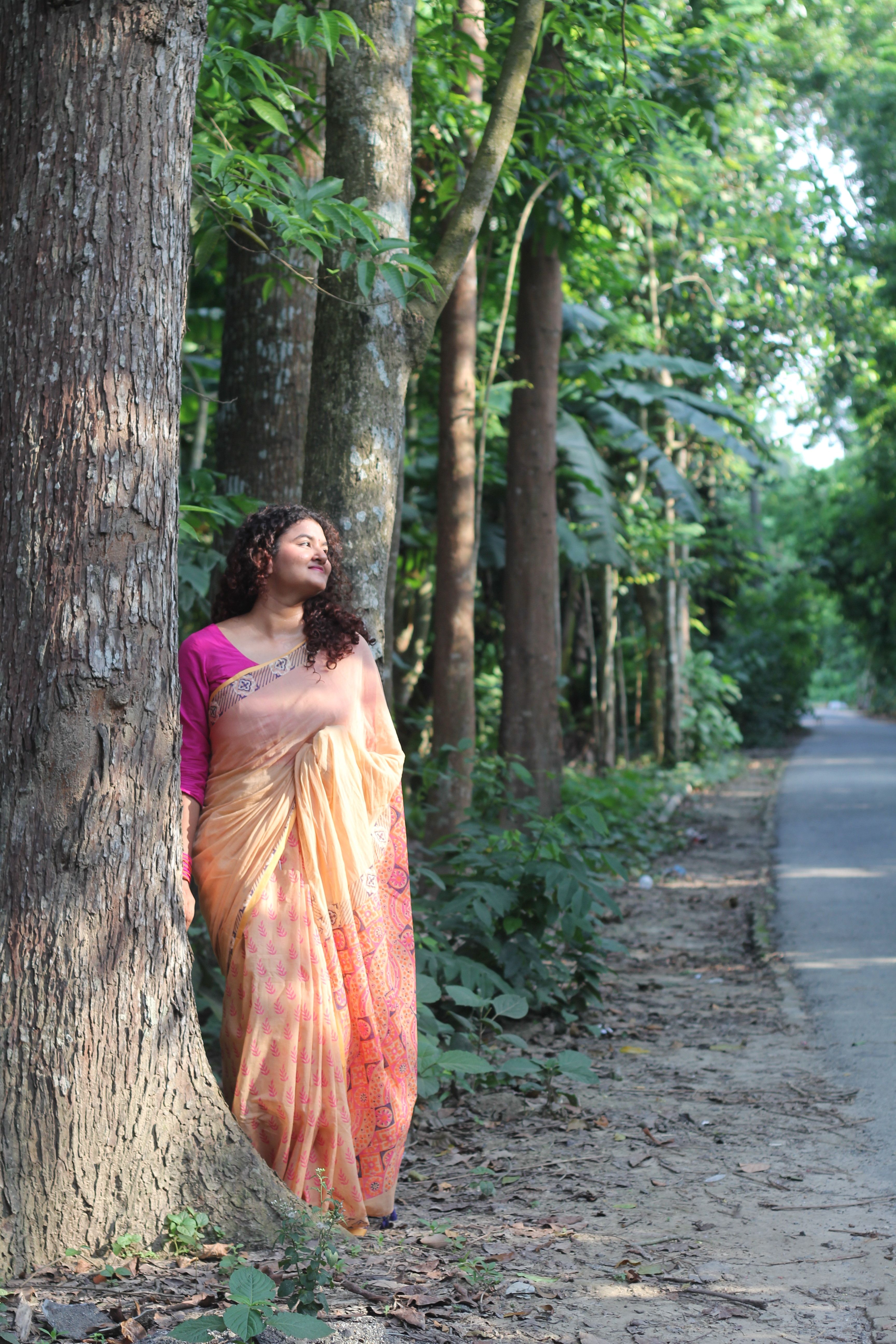 A woman in a saree leans against a tree in a lush forest in Jashore, Bangladesh.