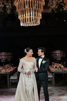 Bride and groom in elegant attire under a stunning chandelier at a wedding reception.