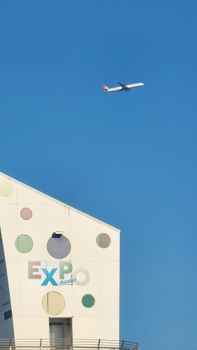 A modern airplane soars above the Expo Antalya building under a clear blue sky.