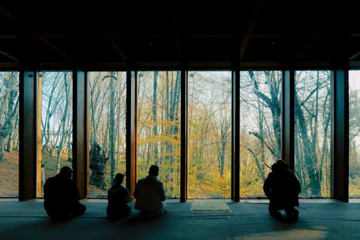 Silhouettes indoors sitting by large windows offering an autumn forest view, serene and contemplative setting.
