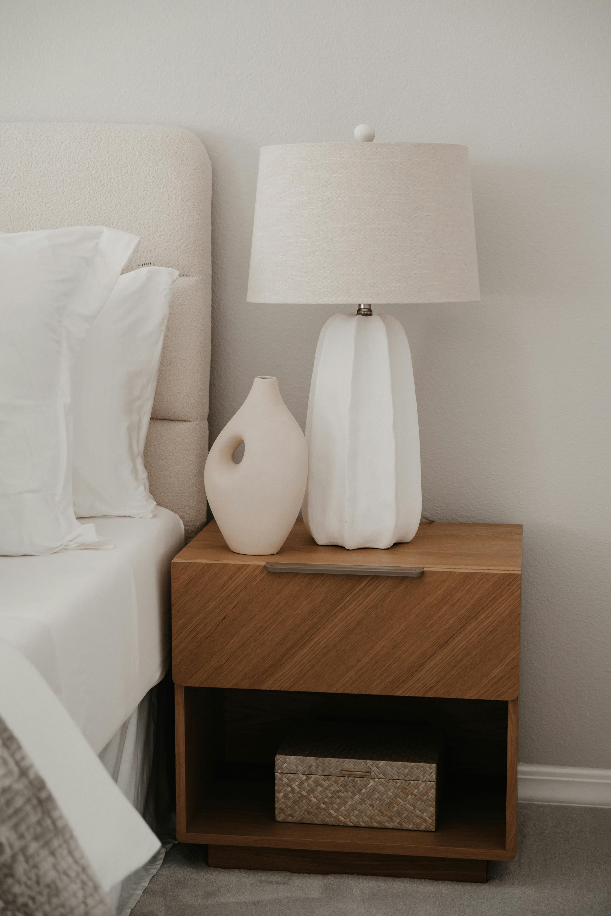 Elegant minimalist nightstand setup with a lamp and vase in a serene bedroom setting.