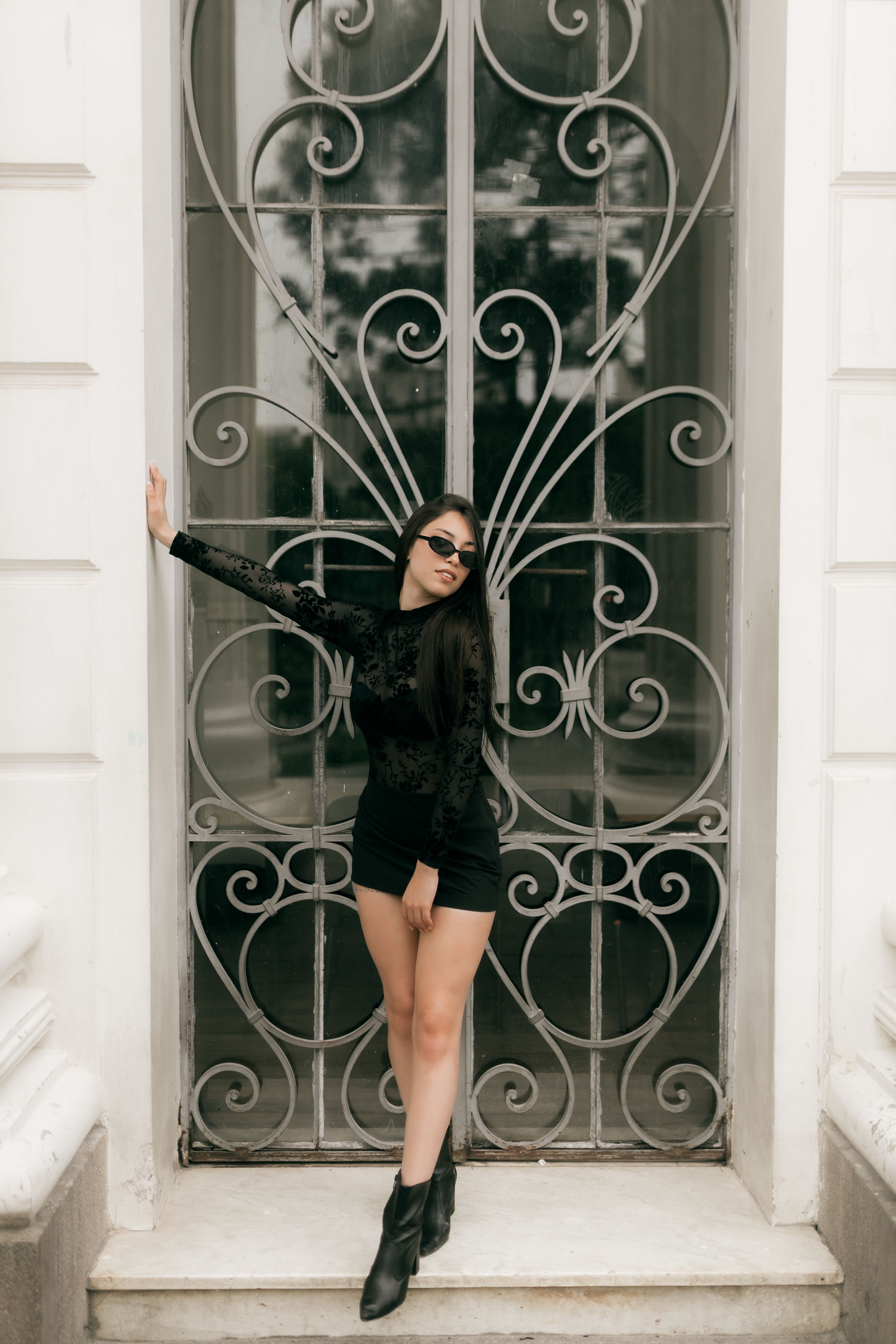Mulher chique em traje preto posa de forma estilosa ao lado de um portão de ferro decorativo ao ar livre.