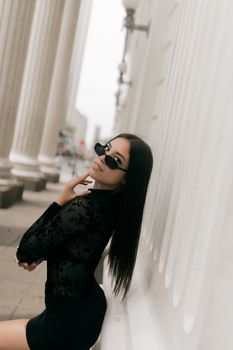 Elegant woman in black dress and sunglasses leaning against white columns.