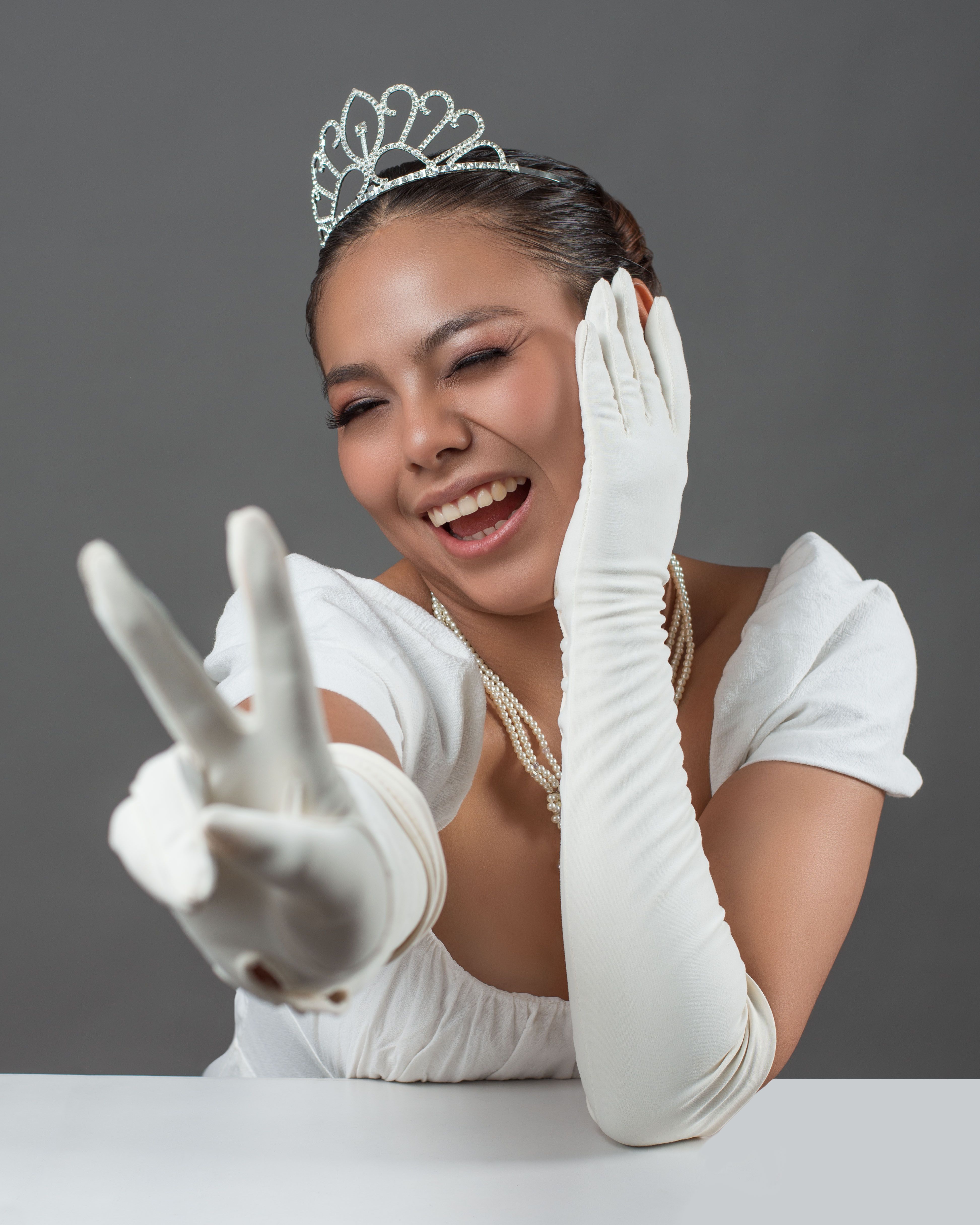 Joyful woman in white gown and tiara making a peace sign, expressing elegance and positivity.