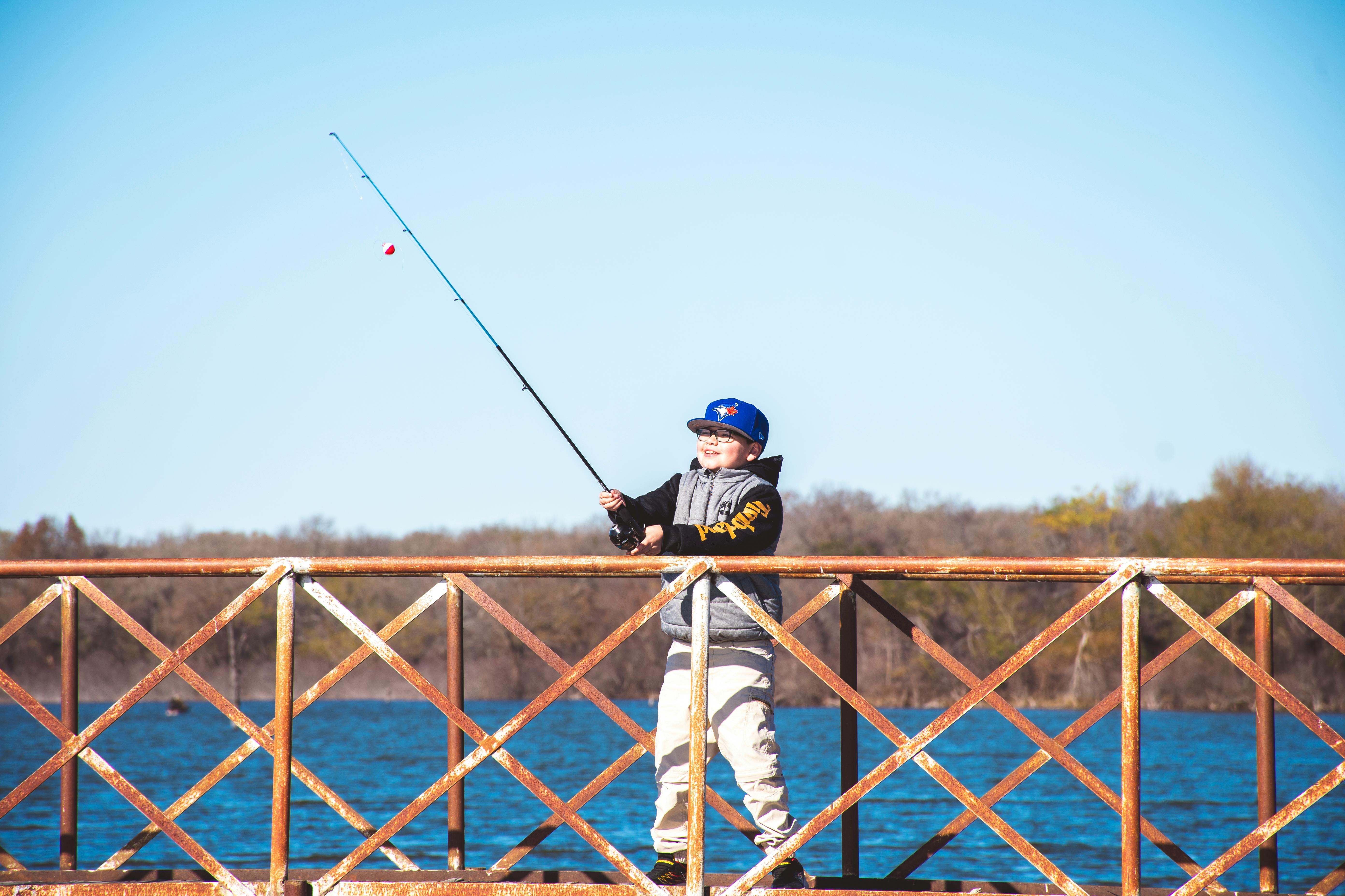 A young boy fishes from a weathered pier on a sunny day, surrounded by water and wooded landscape.