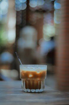 A glass of iced coffee with a straw on a wooden table with bokeh background outdoors.