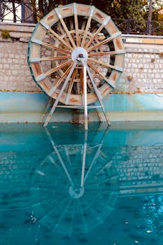 Rustic water wheel reflecting in tranquil blue pool for serene scenic view.