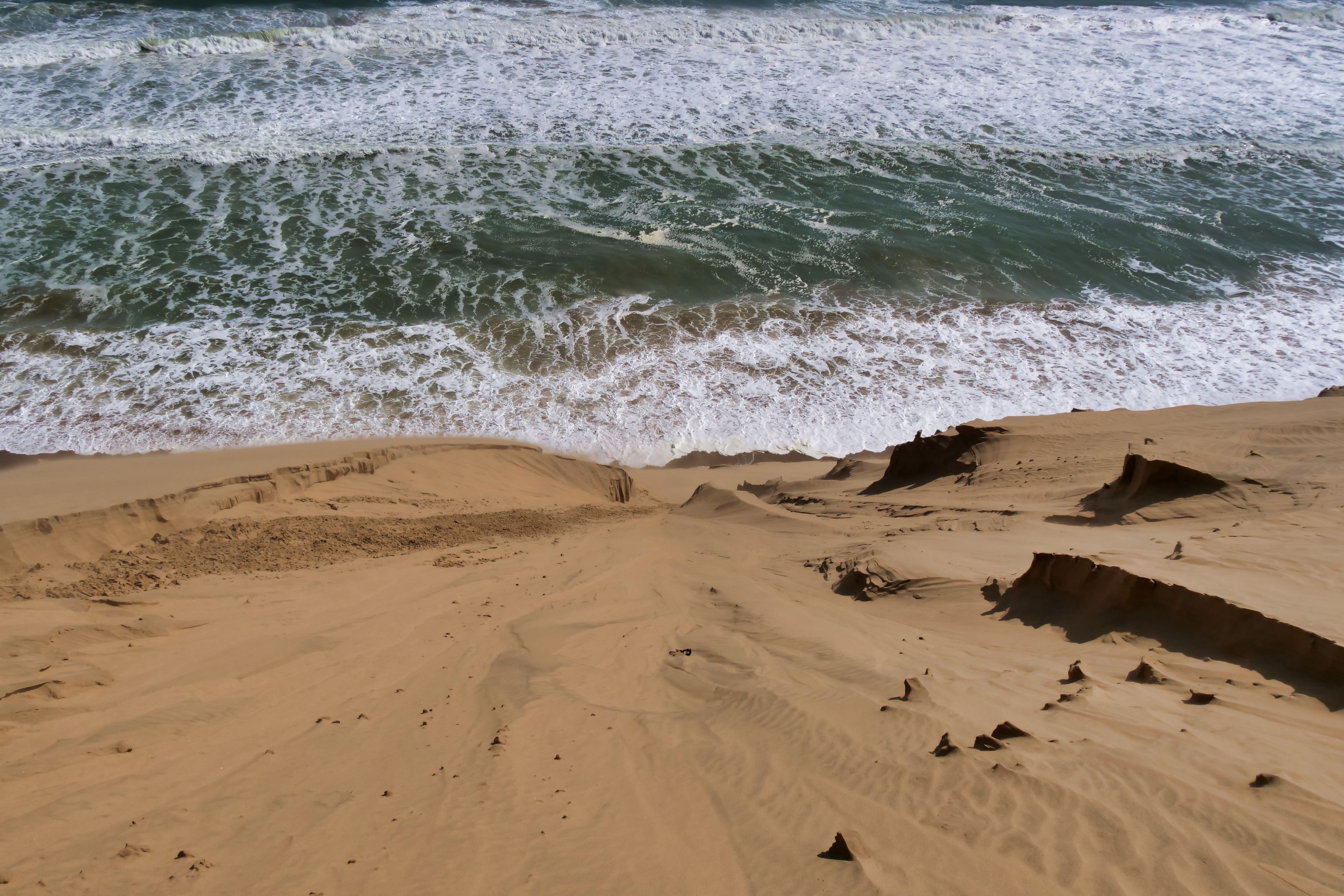 A dramatic landscape of sand dunes cascading into the ocean waves on a sunny day.