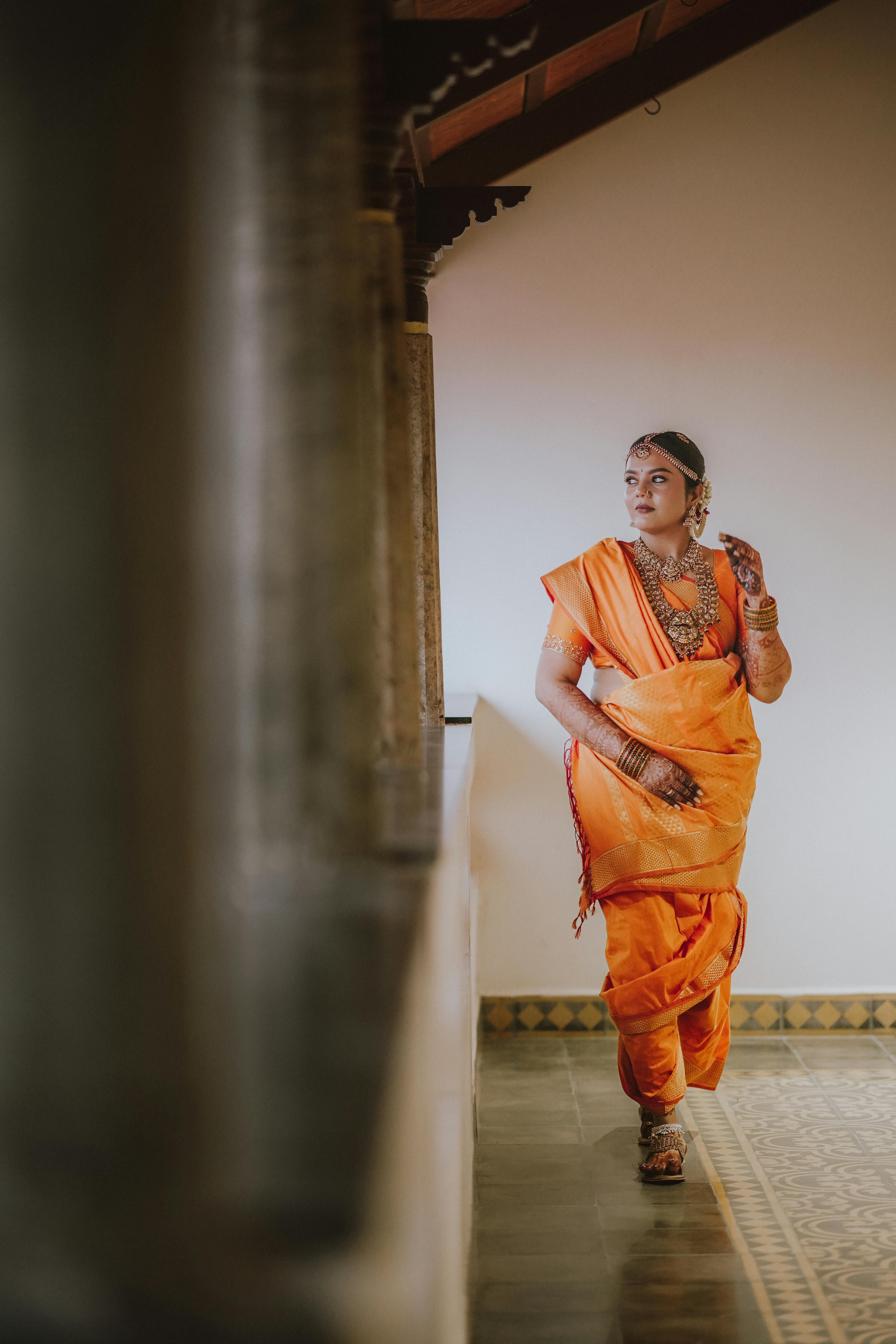 Elegant woman wearing orange saree and jewelry in cultural attire indoors.