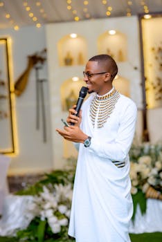 A man in traditional attire speaks into a microphone at a decorated indoor event.
