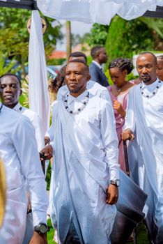 Men in traditional attire participate in an outdoor African wedding procession.