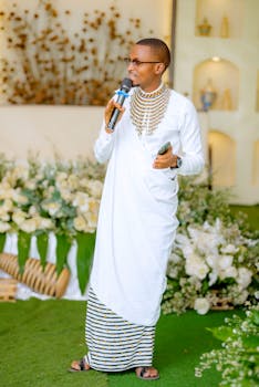 Person in traditional white attire delivers a speech indoors with floral decor.