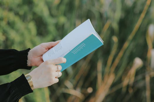 Person in black sleeves holding and reading a book outside among green plants.