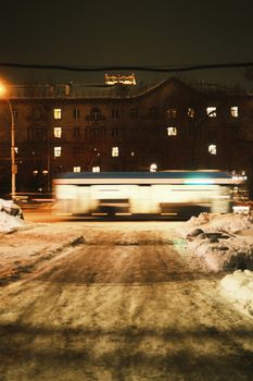 A nighttime winter cityscape in Novosibirsk featuring a blurred bus in motion, against an illuminated building backdrop.