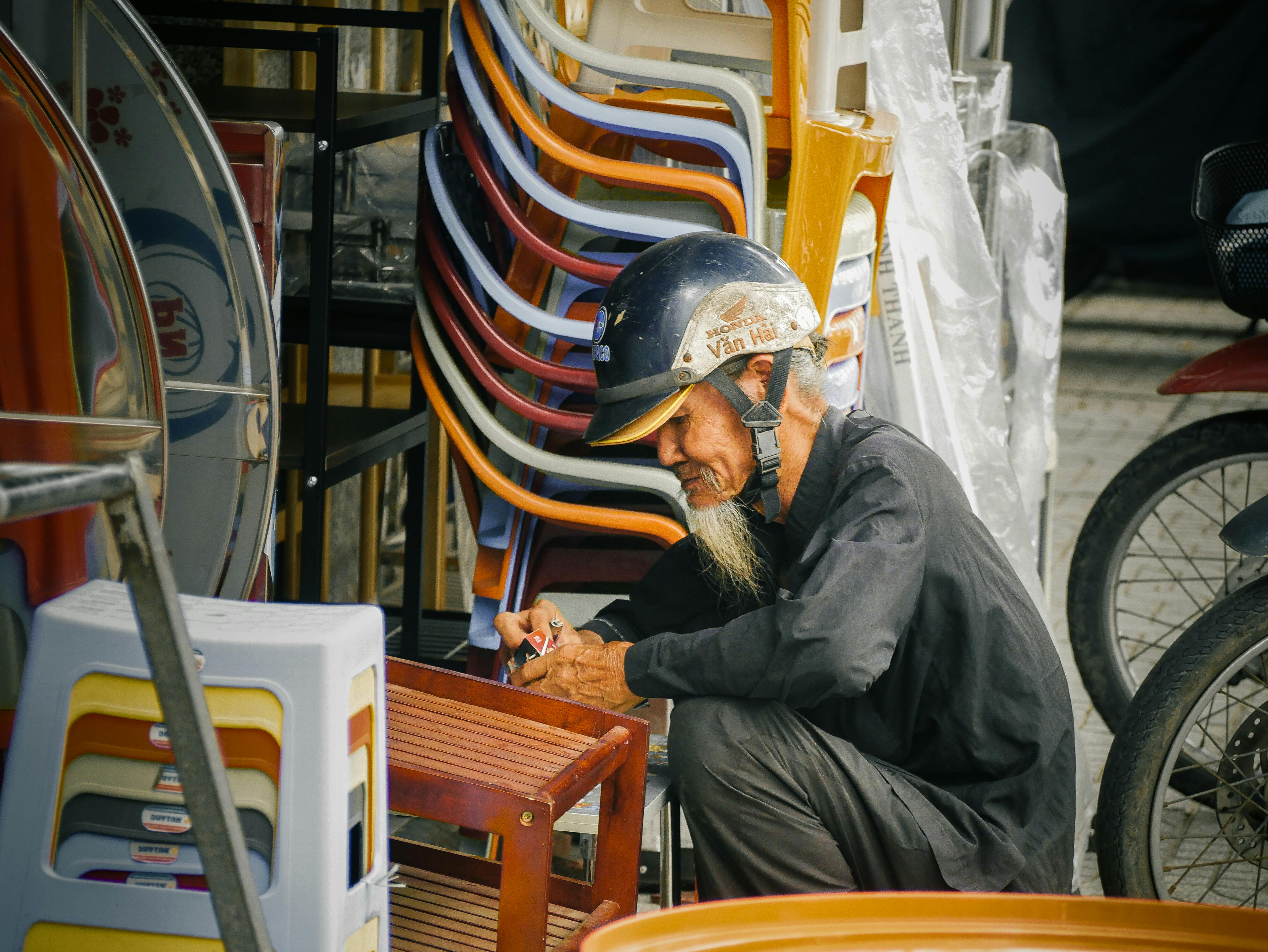 An elderly man wearing a helmet works on furniture repair at an outdoor market, surrounded by colorful chairs.
