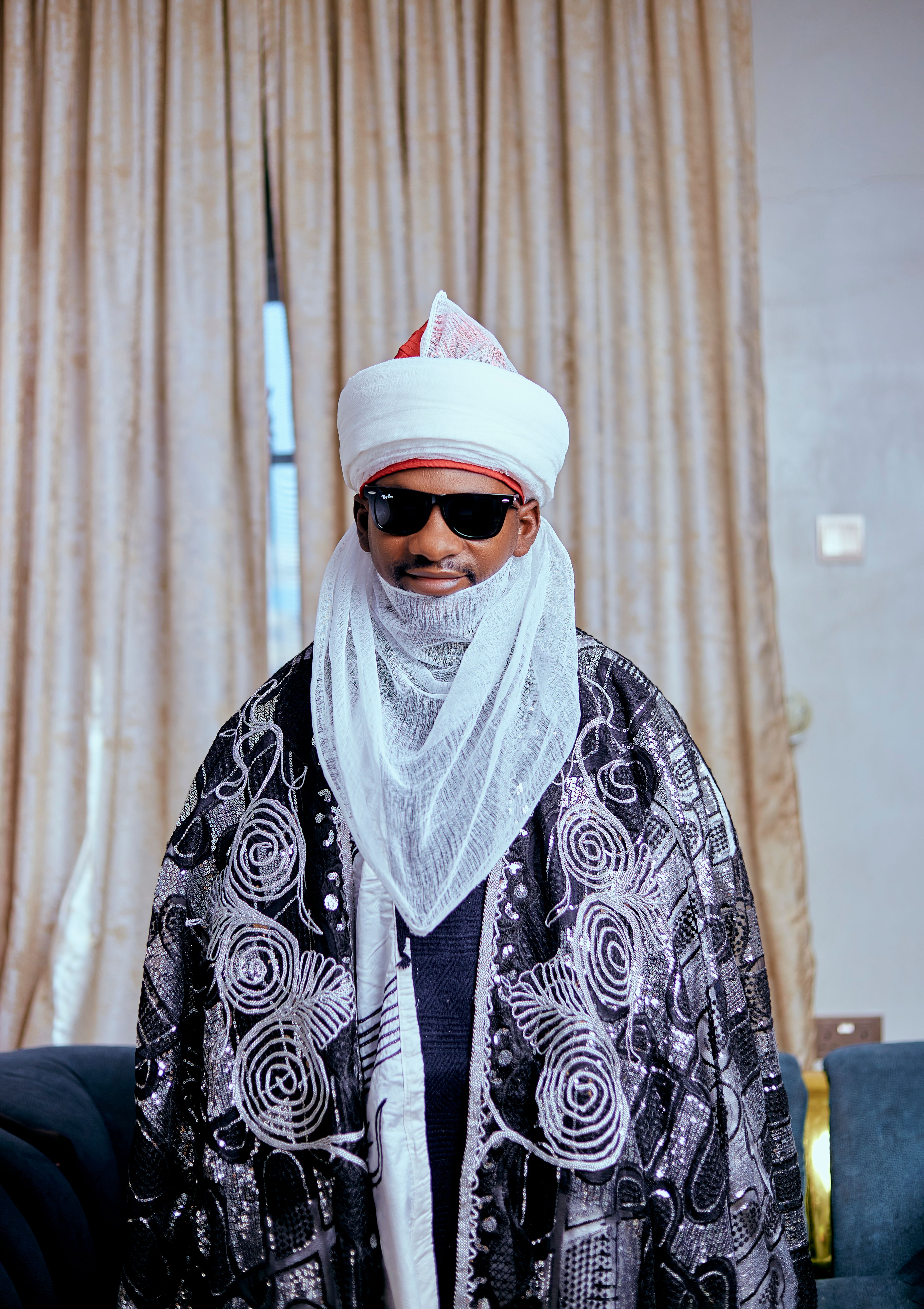 Man in ornate traditional African clothing with turban and sunglasses indoors.