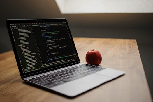 Open laptop displaying code next to a red apple on a wooden desk.