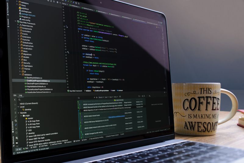 Close-up of laptop with coding software and a motivational coffee mug on a desk.
