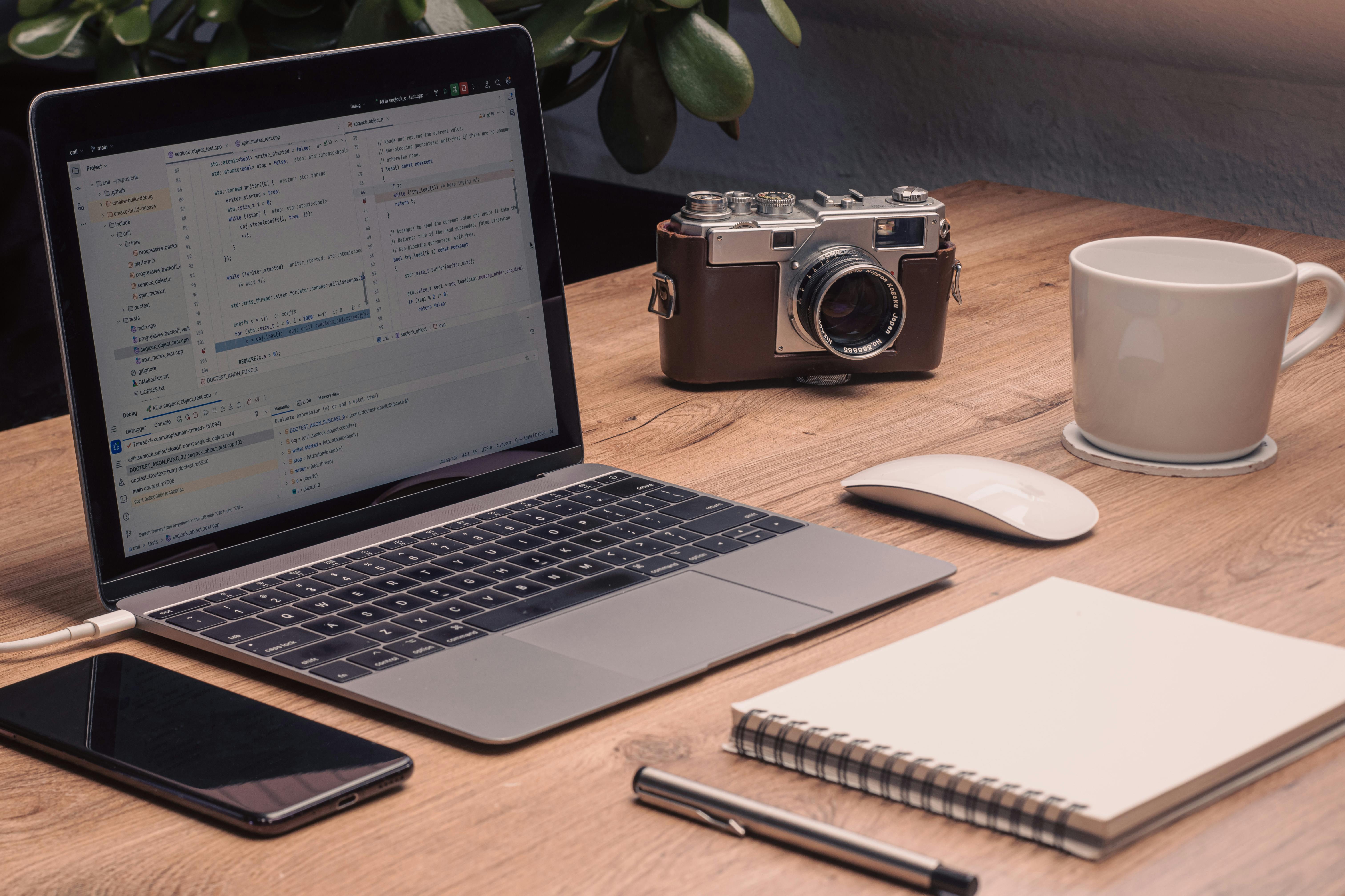A warm, artistic workspace setup featuring a laptop with visible code, vintage camera, and a notebook on a wooden desk.