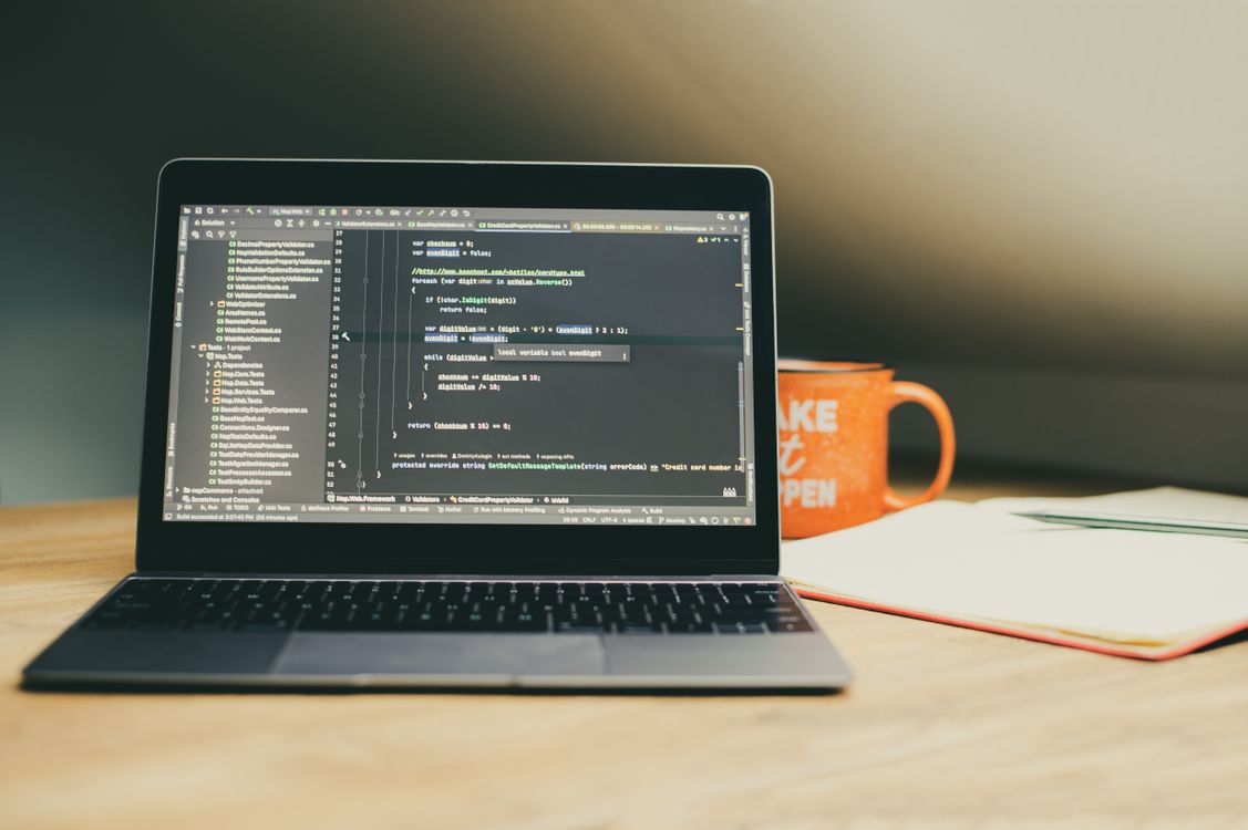 Laptop displaying software code on a desk with a notebook and coffee mug.