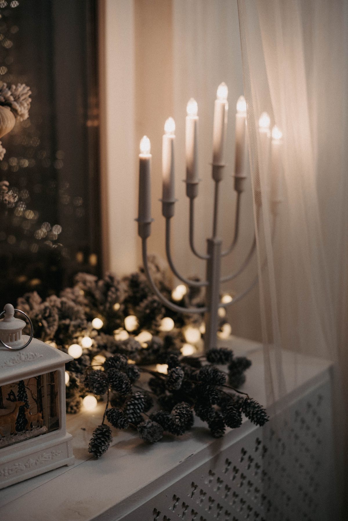 Candle bridge with seven lights on a windowsill beside pinecones, fairy lights and a small lantern