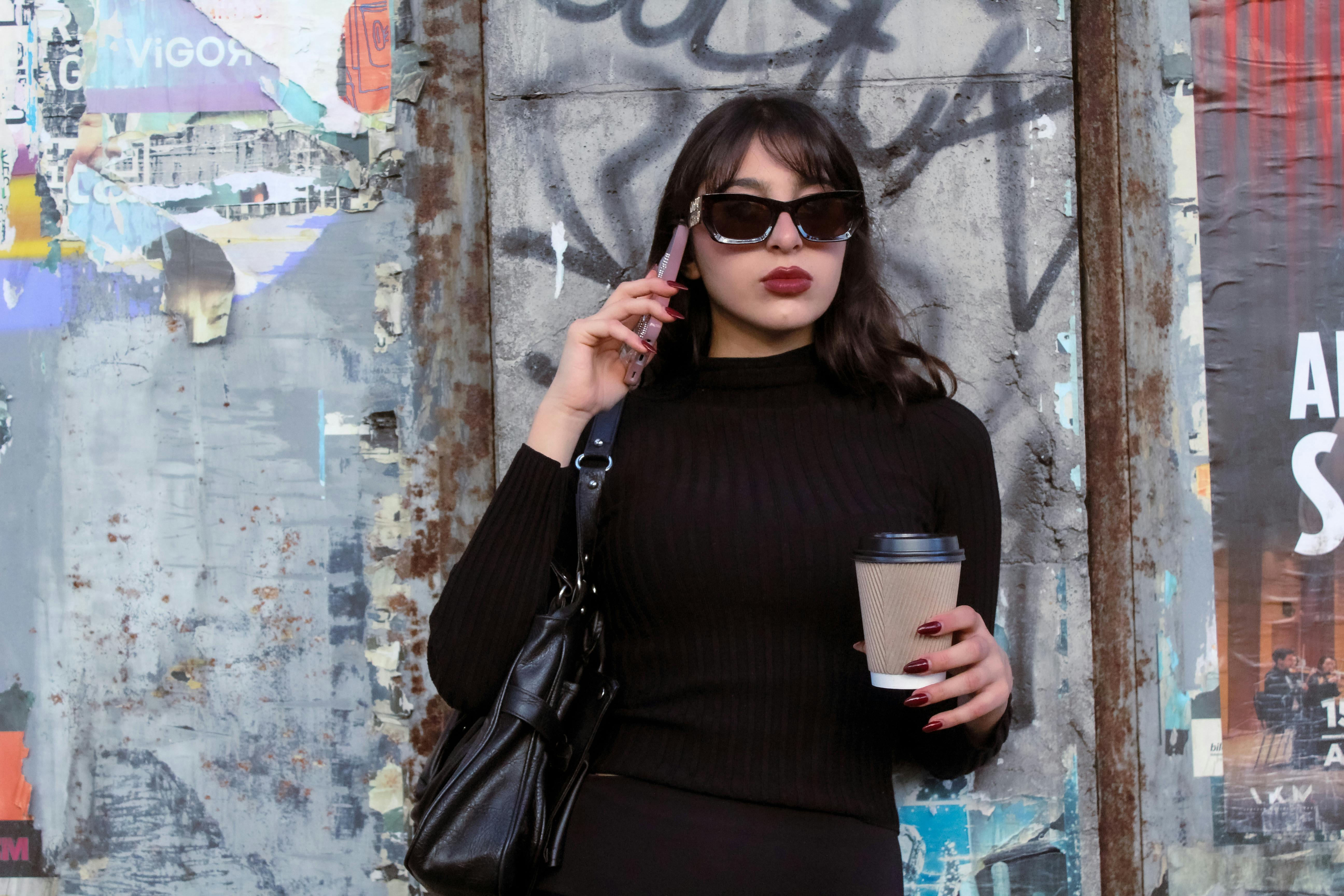 Fashionable woman in sunglasses talks on phone while holding coffee against urban graffiti wall.