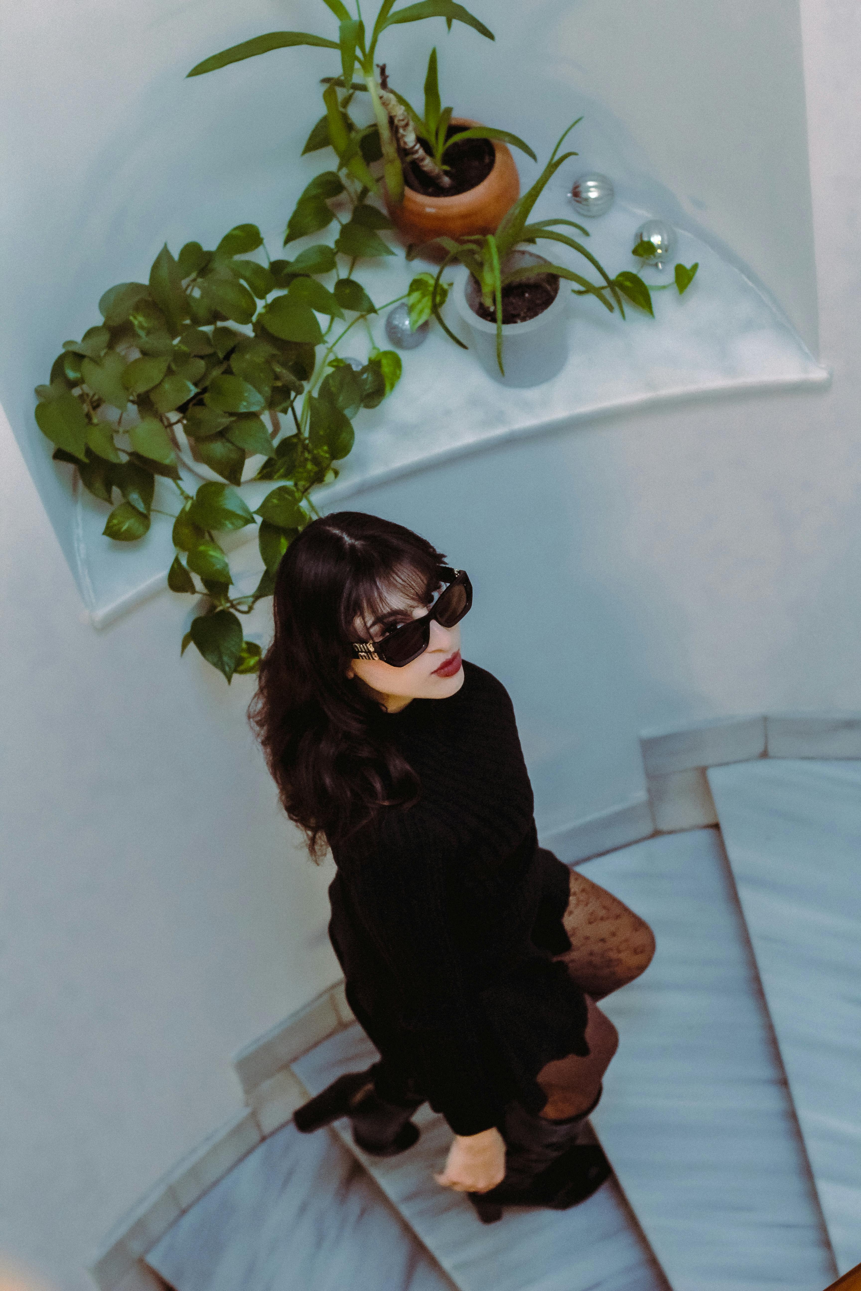 grátis Mulher elegante de óculos escuros em uma escadaria de mármore com plantas, criando um ambiente moderno e sofisticado. Foto profissional