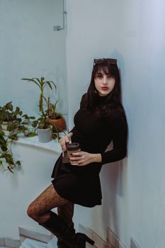 Woman in a black dress leaning against a wall with plants in the background, exuding elegance.