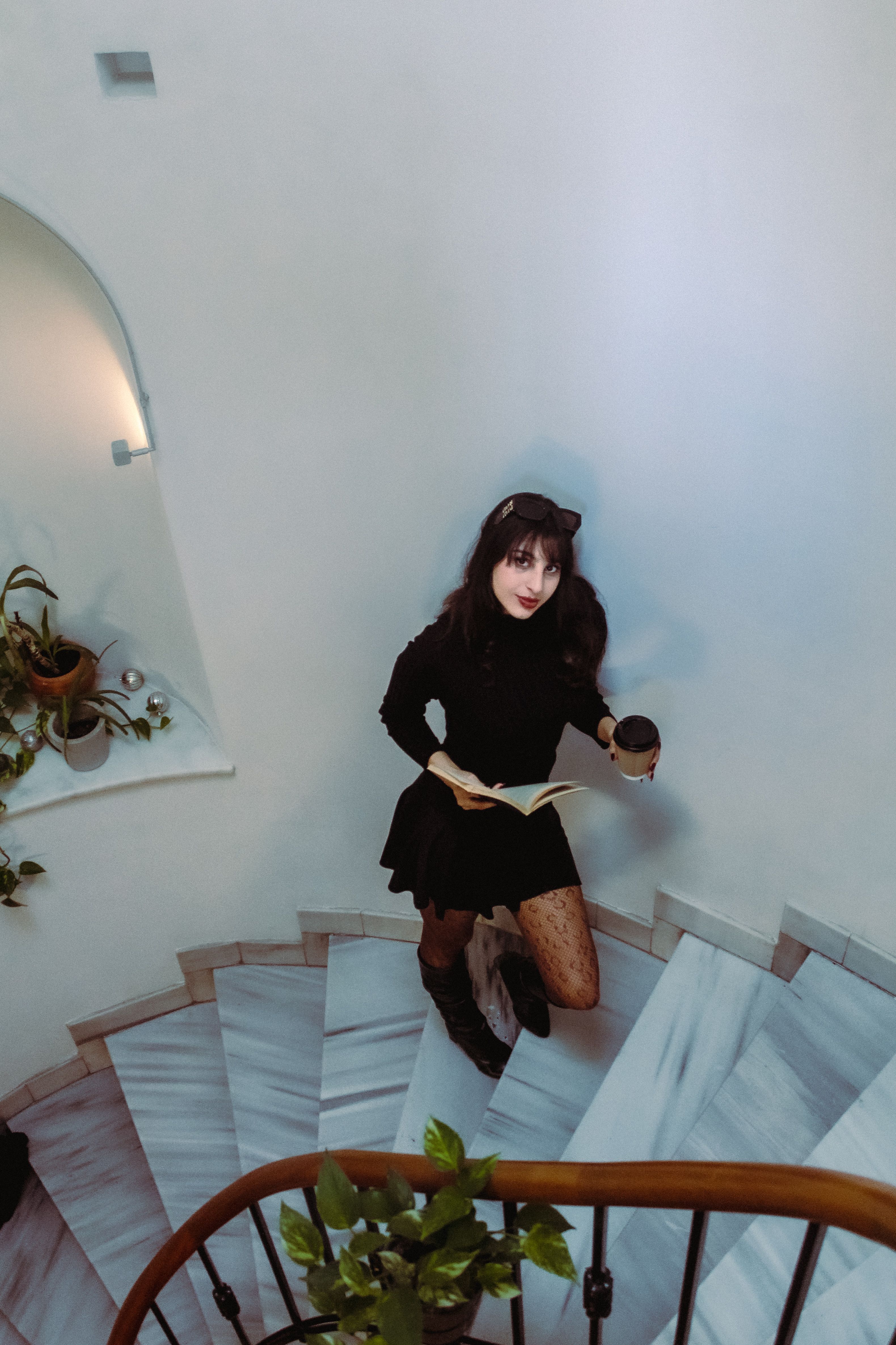 A woman in black holding a book and cup, standing on a spiral staircase indoors.