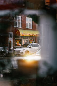 Artistic urban scene of a tattoo shop and a white car reflected in a window.