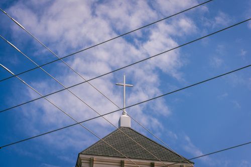 Free A church steeple with a cross set against a blue sky, intersected by power lines, creating an urban religious scene. Stock Photo