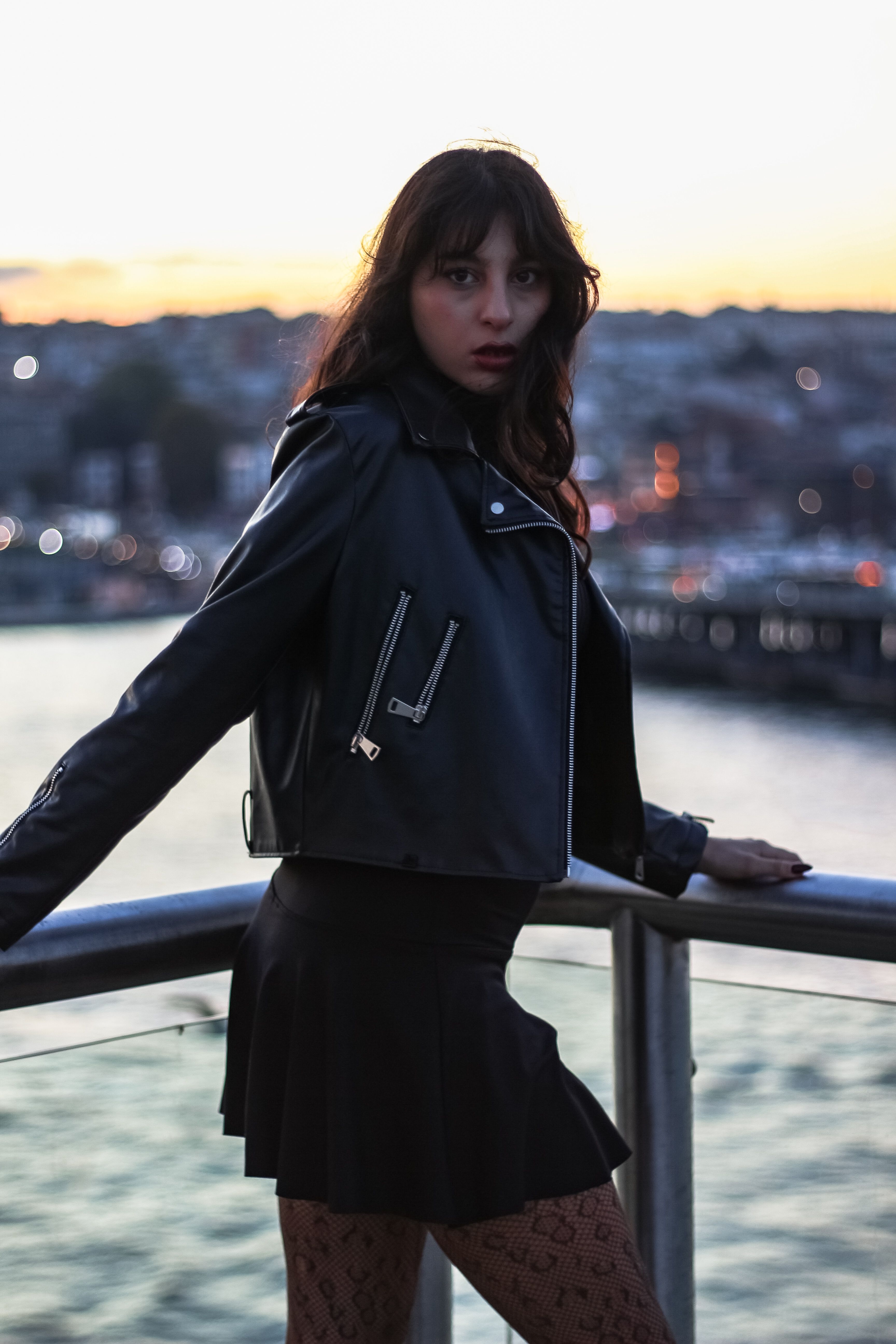 Young woman in a black leather jacket posing near a waterfront during sunset.
