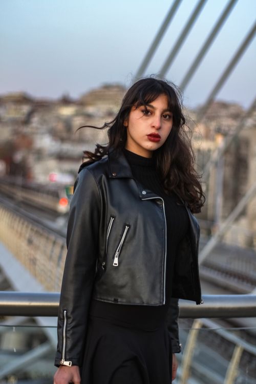 Free Portrait of a young woman in a black leather jacket standing on a bridge with urban background. Stock Photo