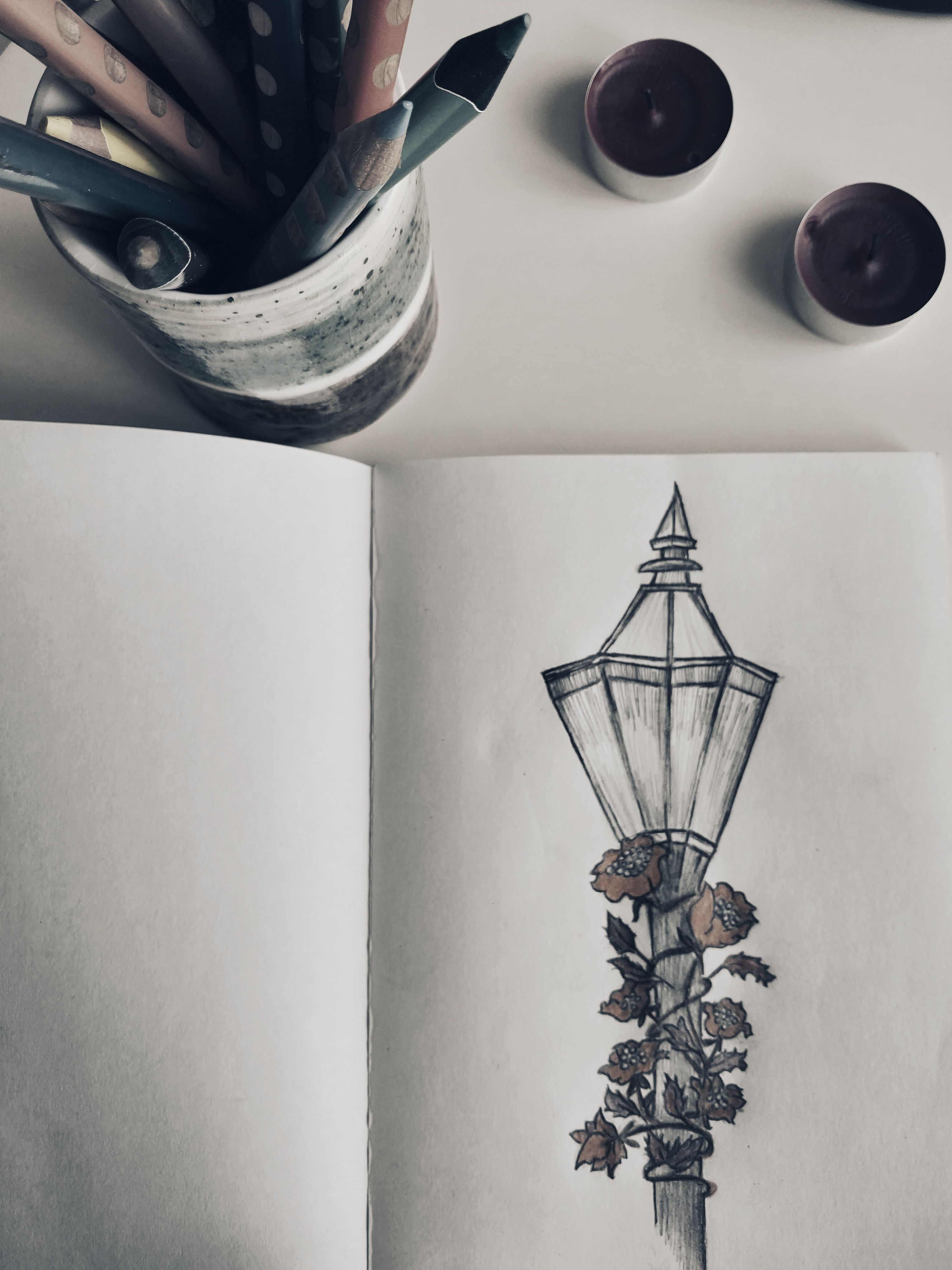 A sketchbook page with a detailed street lamp drawing surrounded by roses, accompanied by colored pencils and candles.