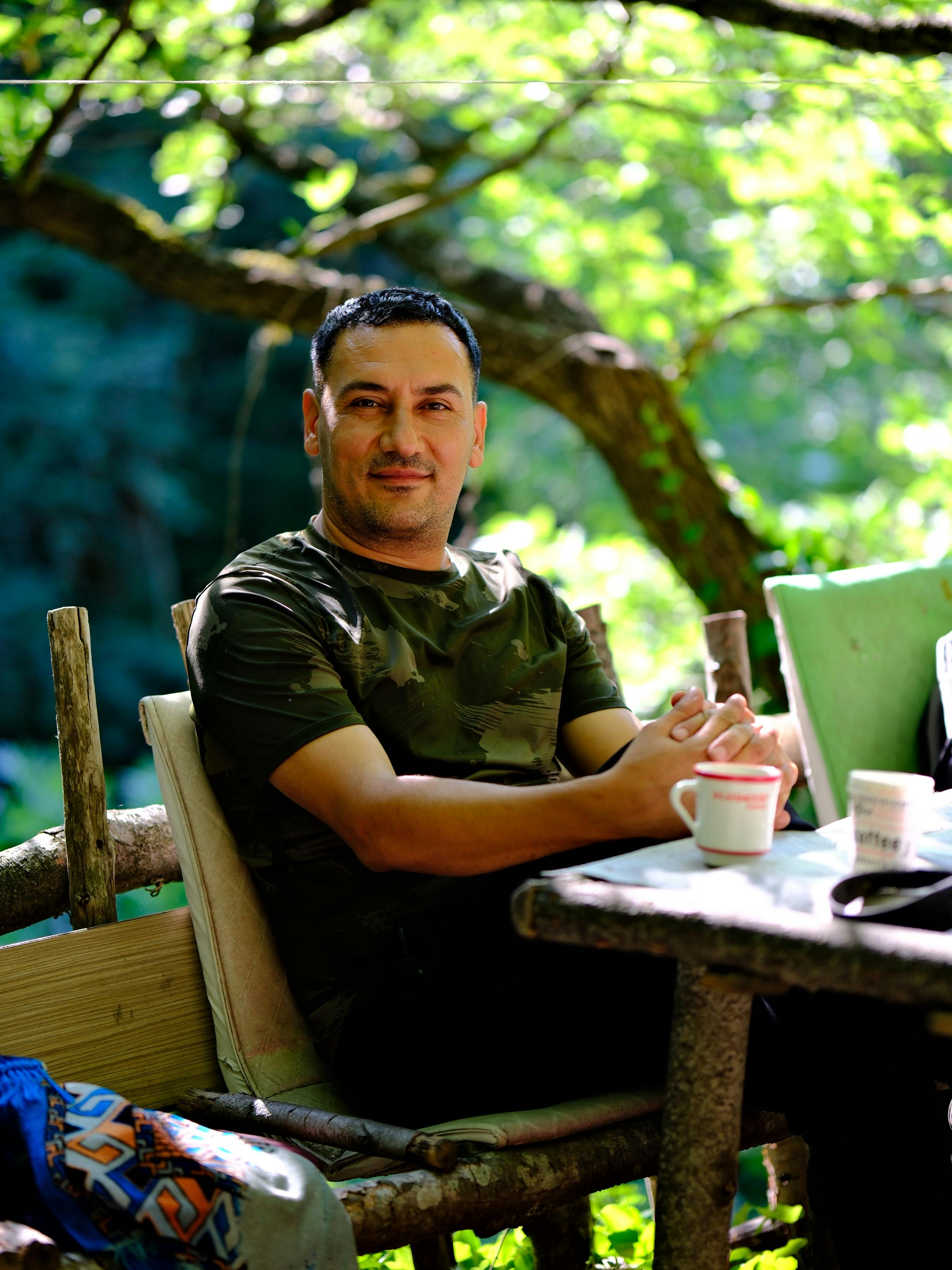Man in camo shirt enjoying coffee at outdoor table surrounded by greenery.