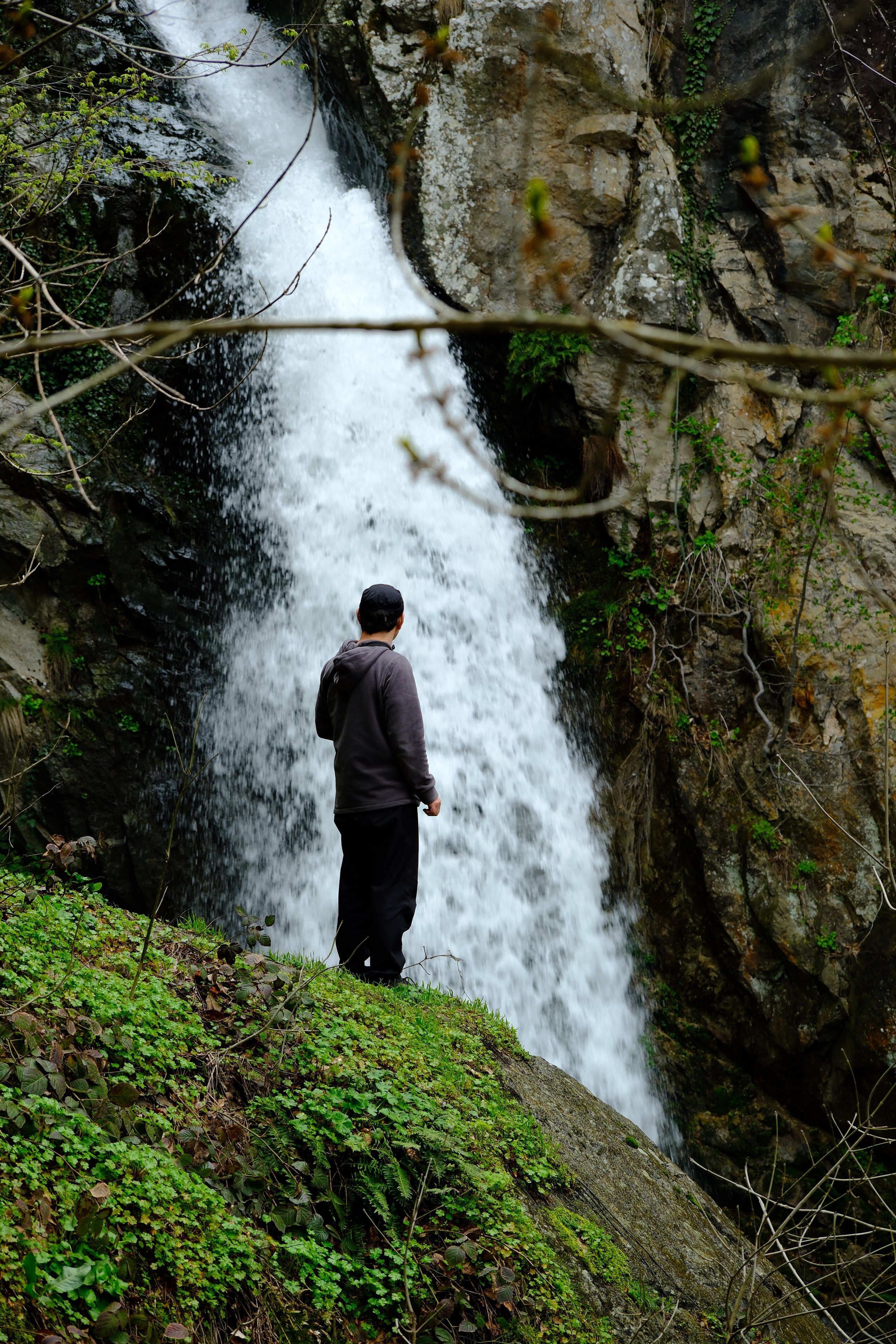 A man stands contemplatively near a powerful waterfall, surrounded by lush nature.
