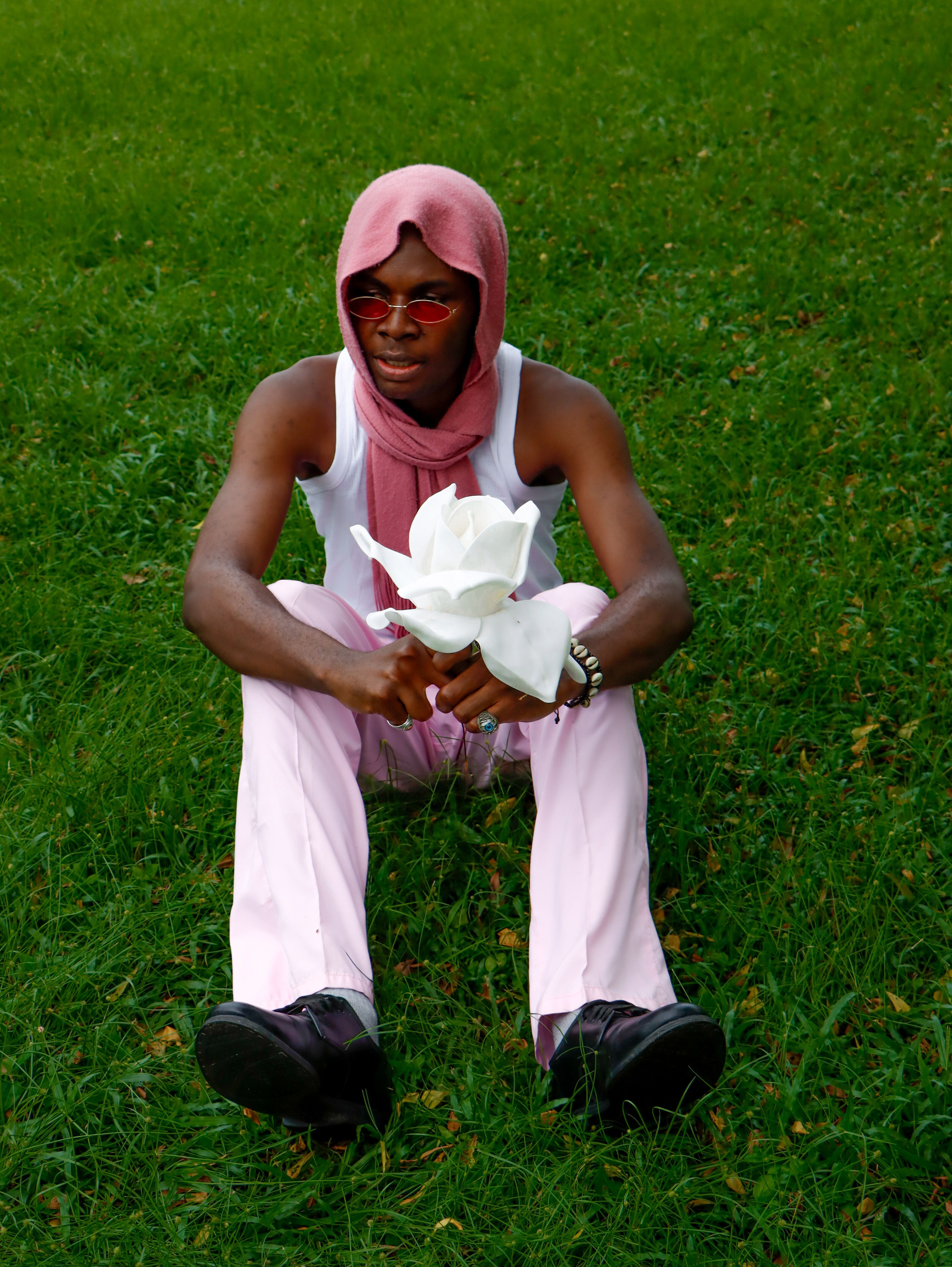 A person in pastel clothing holding a white rose while sitting on grass outdoors.