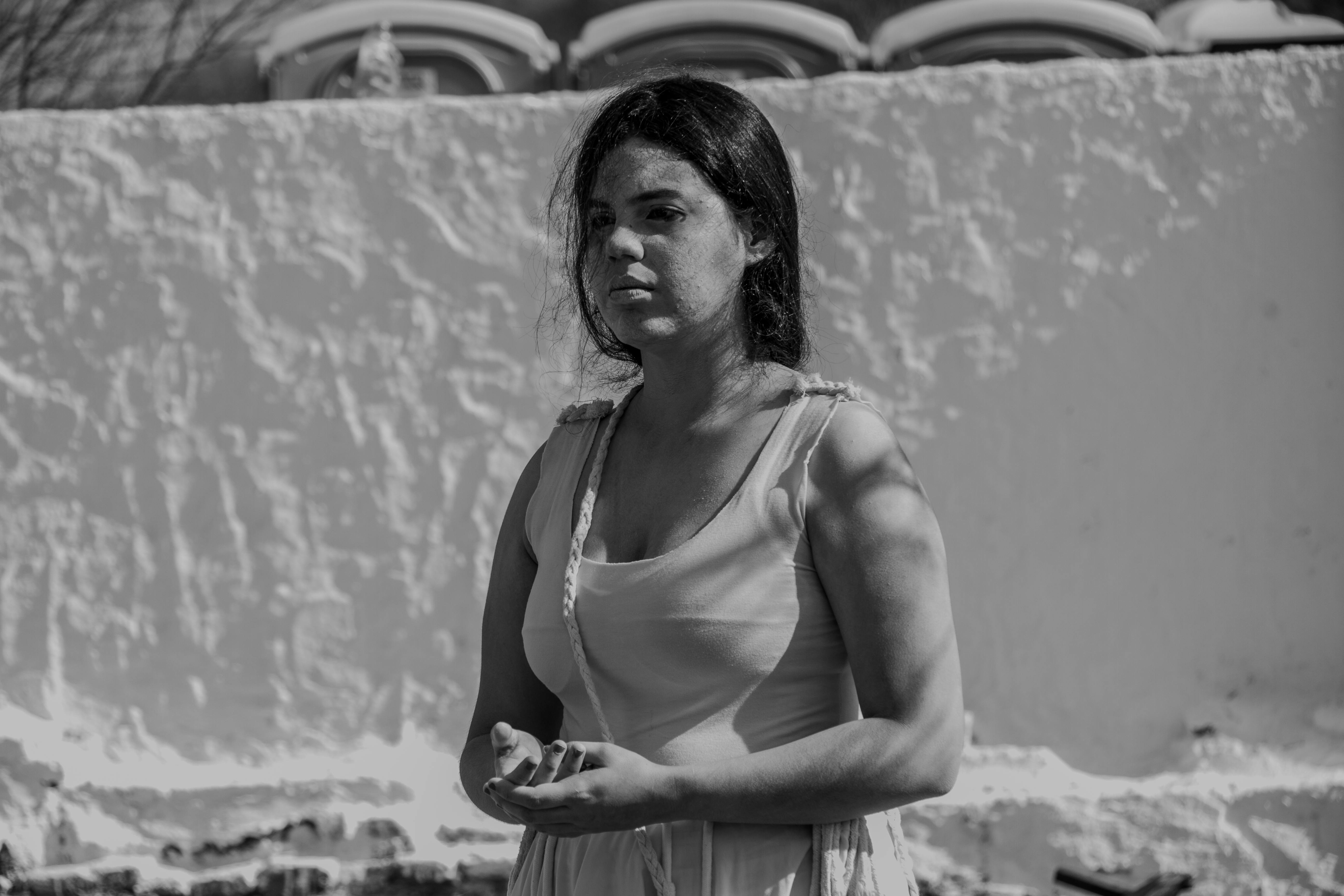 A woman stands thoughtfully in outdoor sunlight, casting shadows on a white wall.