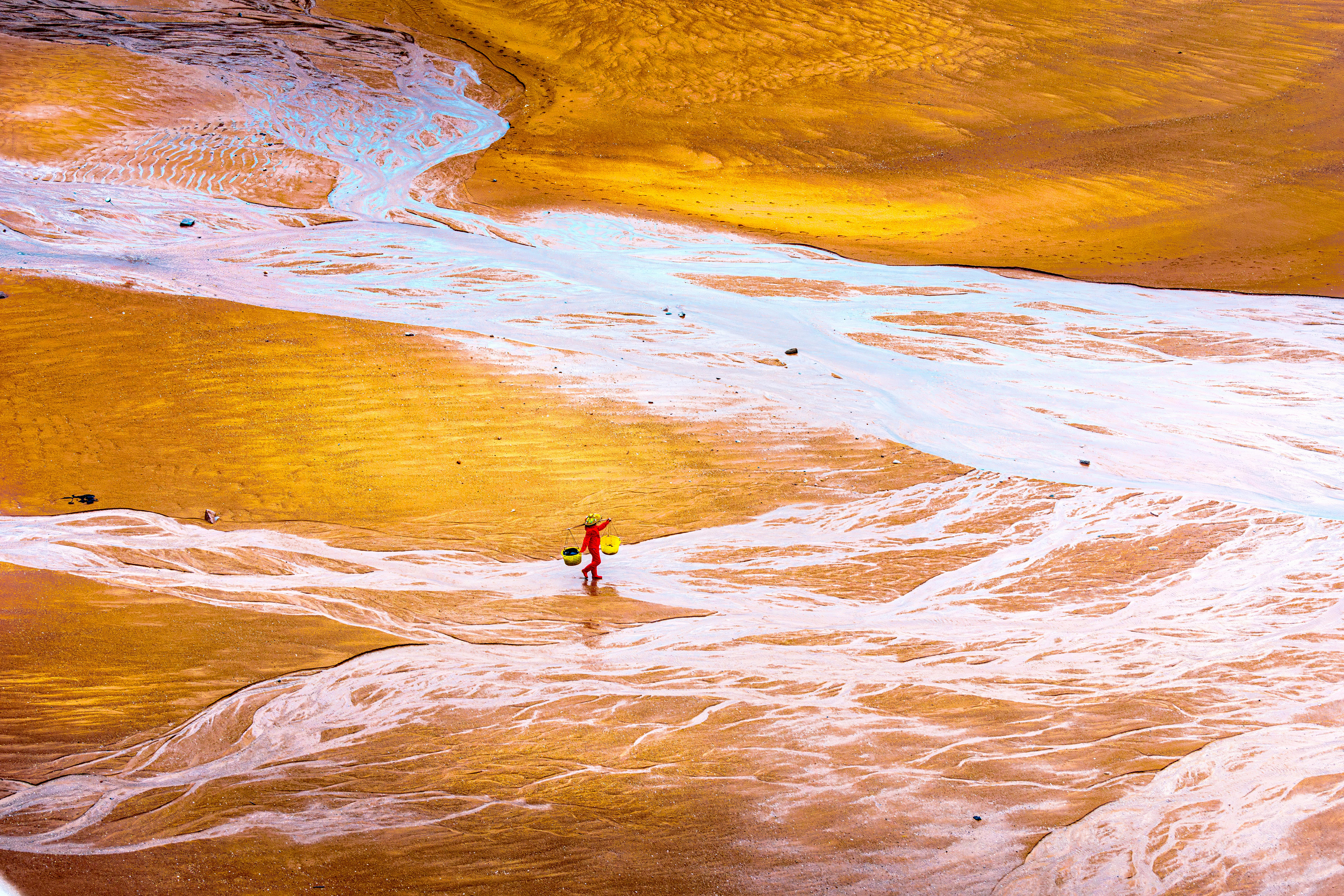 A single person in a hat walking across a colorful tidal flat with striking patterns.