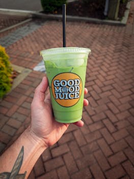 Close-up of a hand holding a green iced beverage with 'Good Mood Juice' label outdoors.
