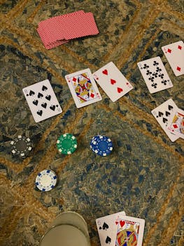 Top view of a poker game scene with scattered playing cards and chips on a patterned carpet.