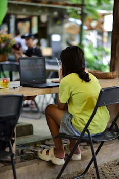 A young woman in casual attire works on her laptop in a cozy outdoor cafe setting.