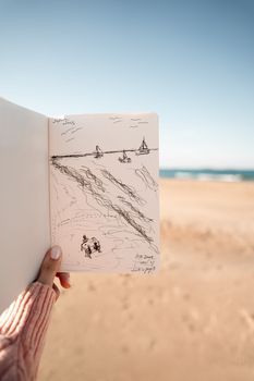 Person holding a sketchbook drawing of the beach scene with blue sky and ocean waves.