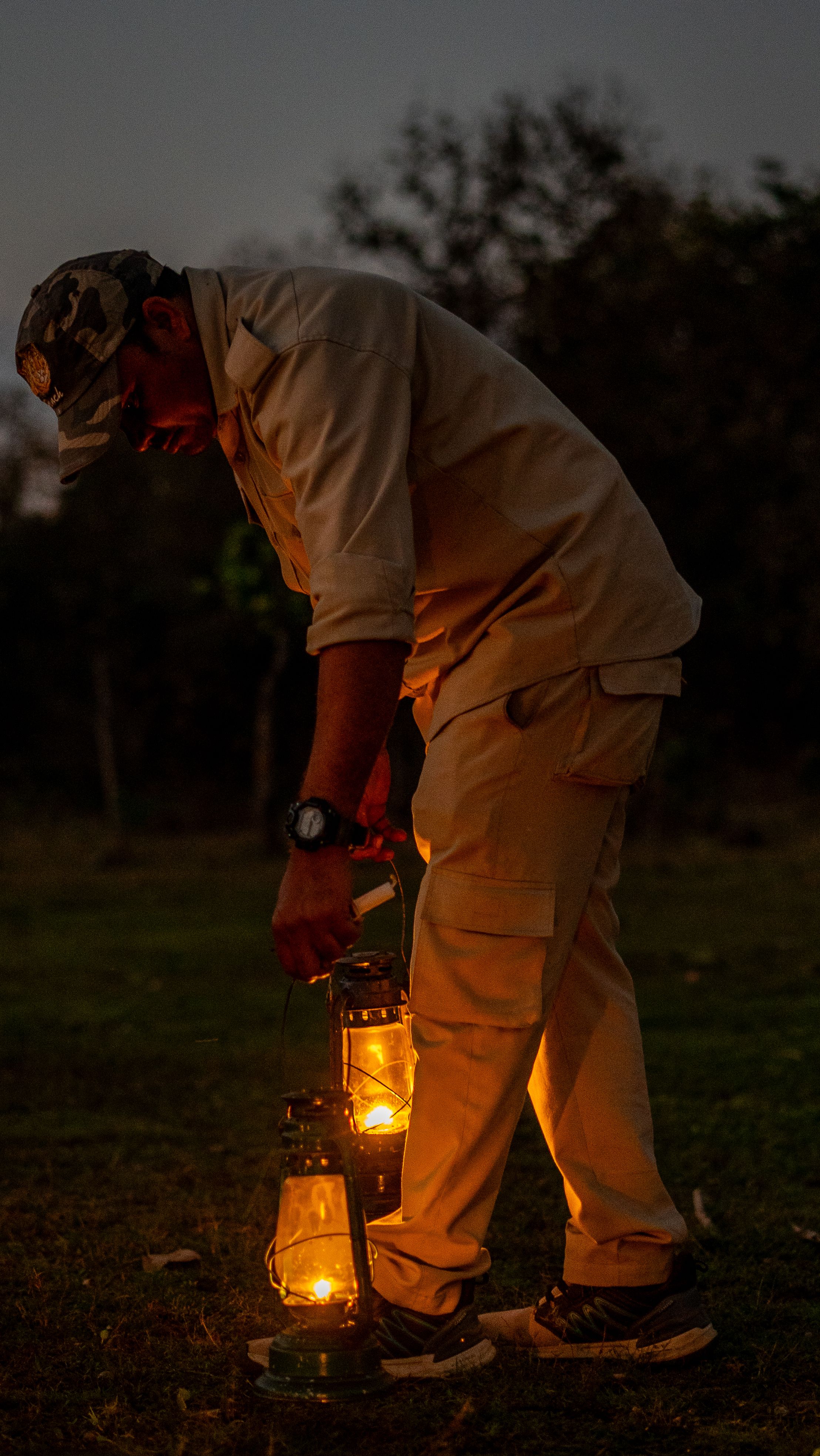 A man in outdoor attire sets up lanterns in a dark forest at twilight, creating a warm ambiance.