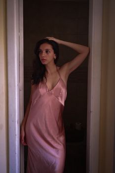 A woman in a pink silk dress poses thoughtfully in a doorway, capturing intimate elegance.