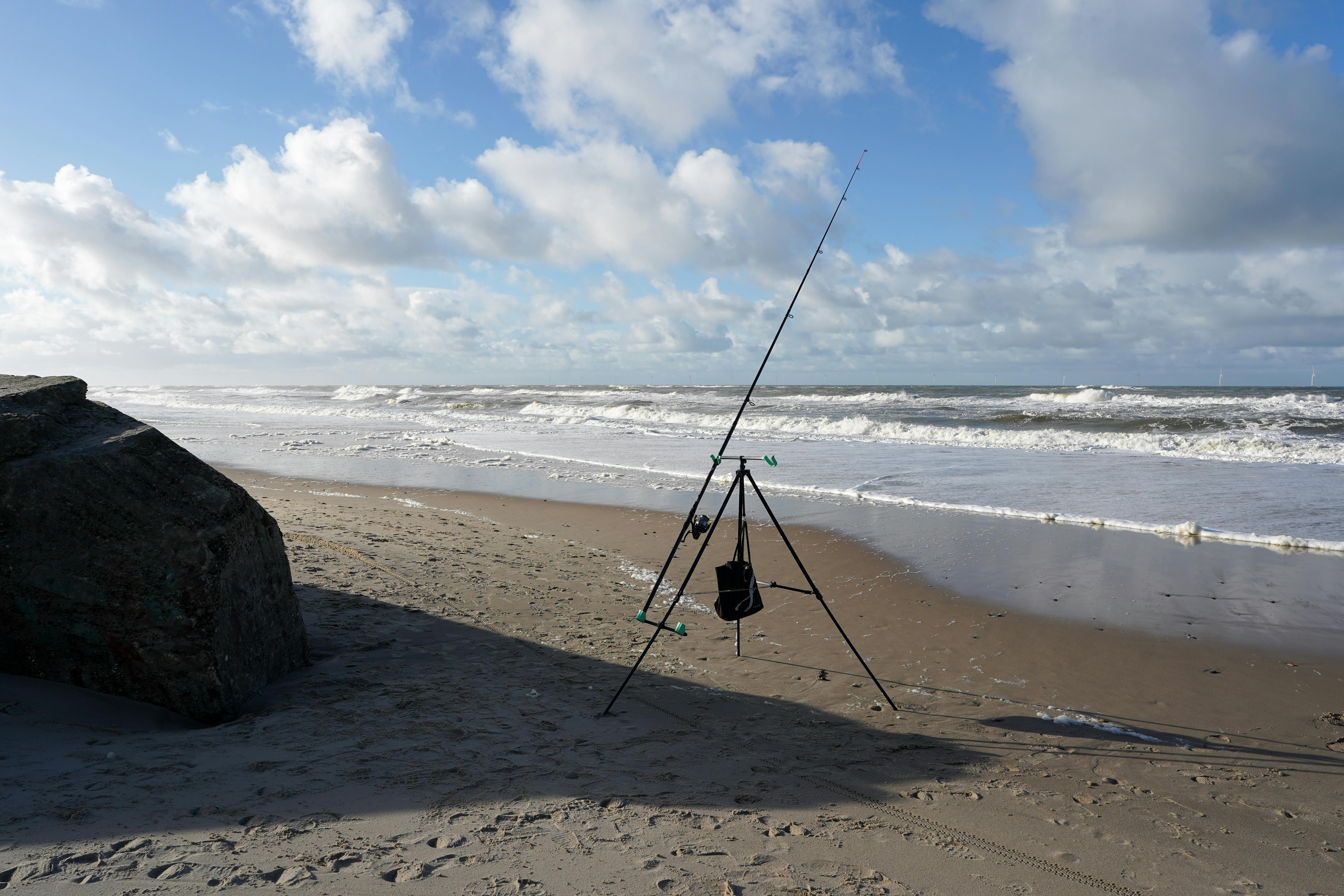 A lone fishing rod stands on a remote beach in Søndervig, Denmark against a backdrop of breaking waves and a vast horizon.