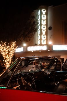 A classic convertible car with Christmas lights parked by an illuminated theater at night.