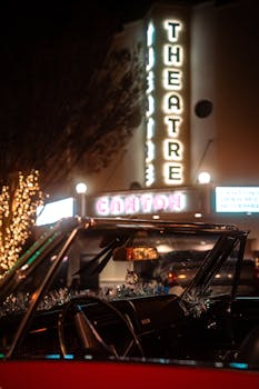 A classic convertible parked outside a lit-up theater at night, with holiday decorations visible.
