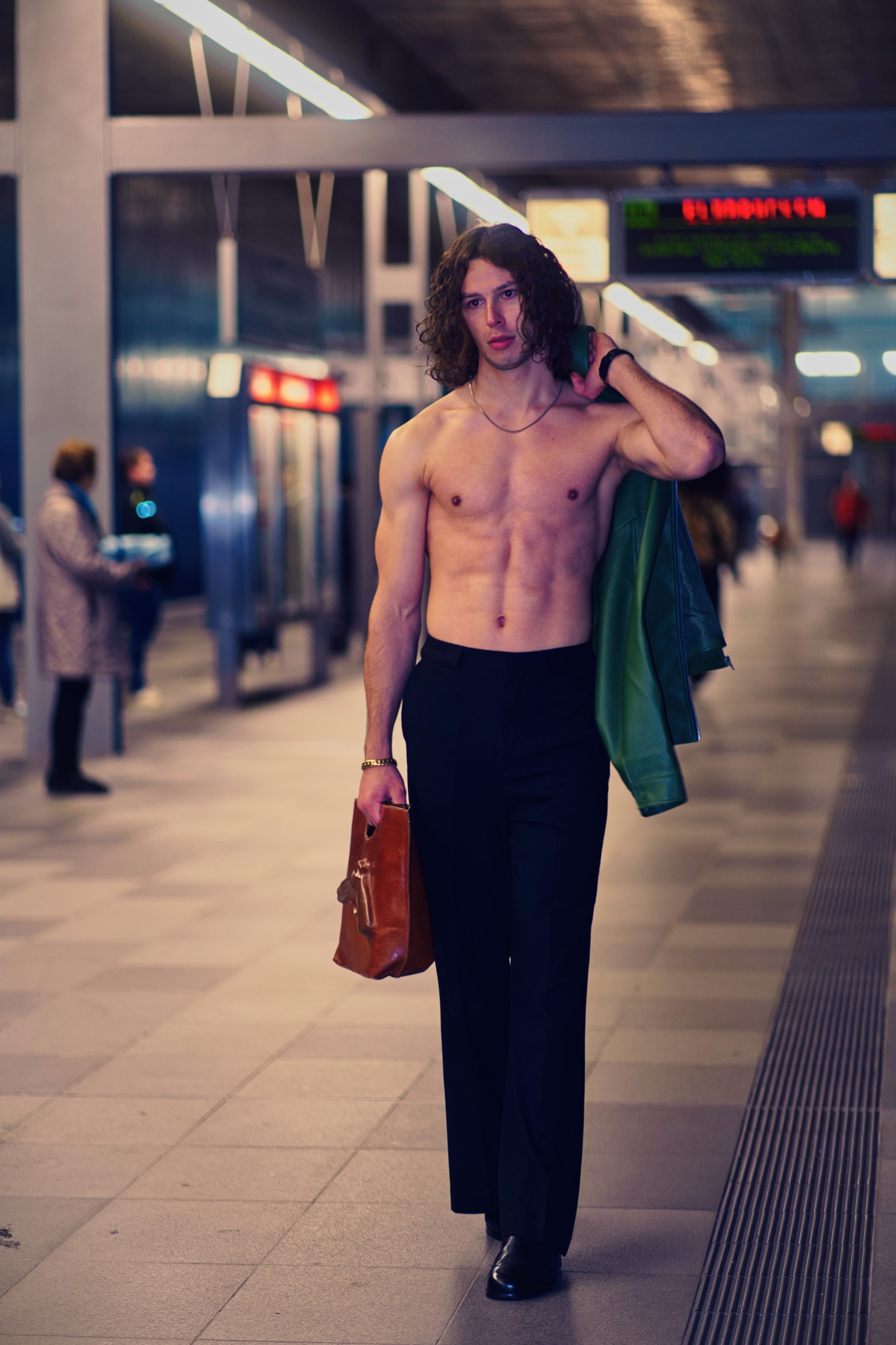 Shirtless man walks boldly through a metropolitan subway station, carrying a leather bag.